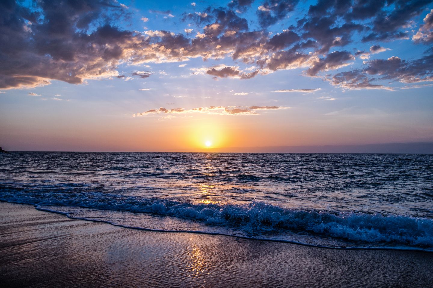 A golden sun setting above the horizon, with blue waves washing onto a sandy beach